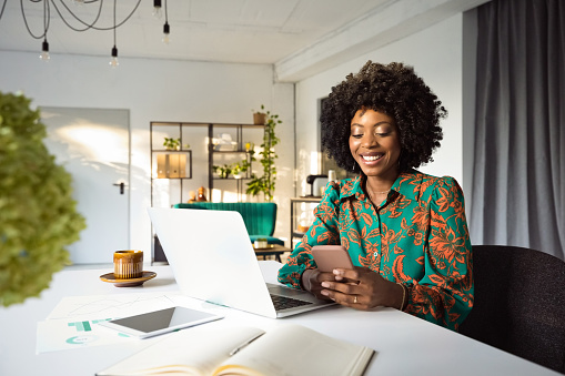 Beautiful, black women wearing floral pattern dress sitting at the table in an office, working on laptop and using smart phone.
