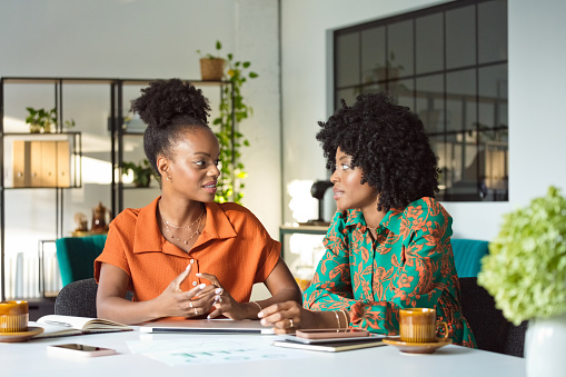 Teamwork concept. Two beautiful, creative black women wearing dresses sitting at the table in an office and discussing.