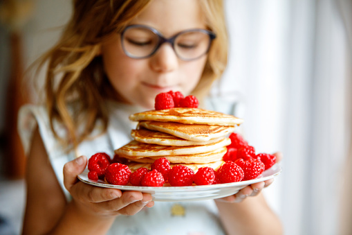 Little happy preschool girl with a large stack of pancakes and raspberries for breakfast. Positive child eating healthy homemade food in the morning