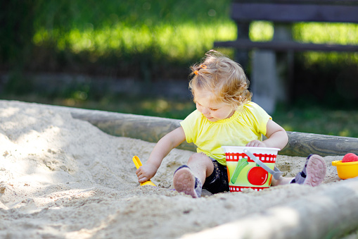 Cute toddler girl playing in sand on outdoor playground. Beautiful baby having fun on sunny warm summer sunny day. Happy healthy child with sand toys and in colorful fashion clothes