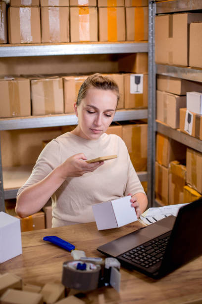 istock Female small business owner using mobile app on smartphone checking parcel box. Warehouse worker, seller holding phone scanning retail dropshipping box postal parcel bar code on cell technology. 1954672804
