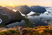 Sunset casts radiant light over Reinebringen, with the sun mirroring on calm fjord waters, the moss-covered foreground leading to Reine's idyllic fishing village