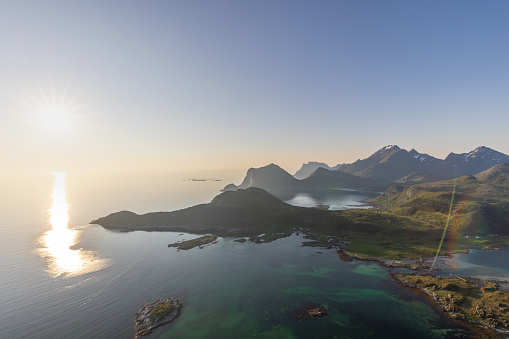 The midnight sun hovers over Offersoykammen, casting a shimmering path across the Nappstraumen, with the Lofoten Islands' peaks rising majestically in the distance