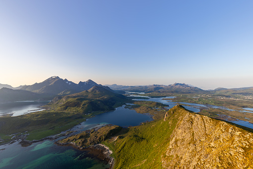 The midnight sun casts a golden glow on Offersoykammen, with the vivid blues and greens of the Nappstraumen area unfolding below. Lofoten Island, Norway