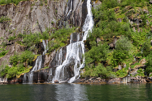 Sunlit water gushes down a steep rocky face into the tranquil waters of Trollfjorden in Lofoten, Norway, framed by lush greenery