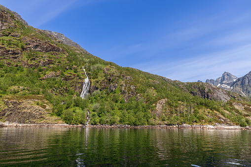Crystal waters tumble from a high cliff into the calm fjord, surrounded by the vibrant greens of a Norwegian summer in Trollfjorden, Lofoten