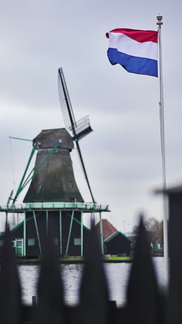 Traditional windmill and dutch flag in Zaanse Schans in the Netherlands, dutch flag with windmills in the Netherlands, ancient wooden windmills in Zaanse Schans, most popular tourist attraction in the Netherlands, dutch national flag