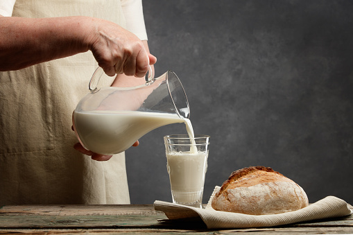 Hands of an elderly woman with a jug of milk. Milk pouring into a glass from milk jug. A loaf of bread and milk on a wooden table. Selective focus