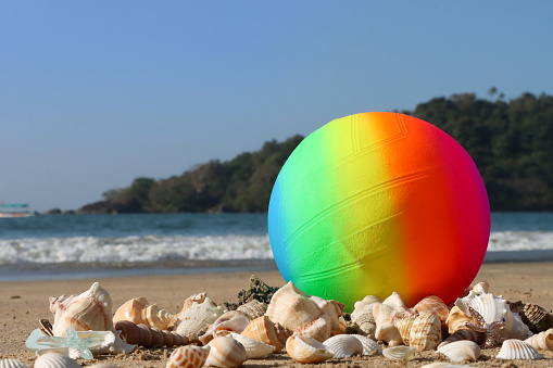 Stock photo showing close-up view of sandy beach with a multicoloured, rainbow striped football on pile of seashells by water's edge of sea at low tide in the background. LGBTQ+ gay holiday concept.