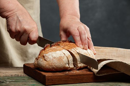 Hands of an elderly woman cutting bread on a wooden table and kitchen board. Selective focus