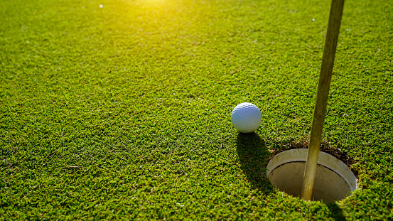 Top view of a golf ball on green course at hole. Golf ball and golf club on green in the evening golf course with sunshine.