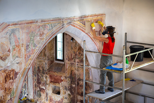 Restoration Process on a Church Arch Wall by Expert Adult Woman Restorer Wearing a Protective Gas Mask While Bringing Out Original Fresco Colors With Use of Some Chemical Treatments