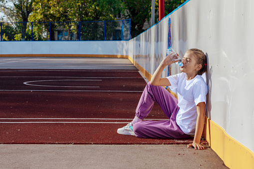 girl drinks water while sitting on a rubber coating on the basketball court. Sport, active lifestyle concept