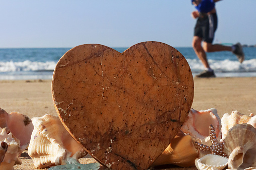 Stock photo showing close-up view of a pile of seashells with a starfish surrounding a sandstone heart standing up in the sand on a sunny, golden beach with sea at low tide in the background. Romantic holiday and honeymoon concept.