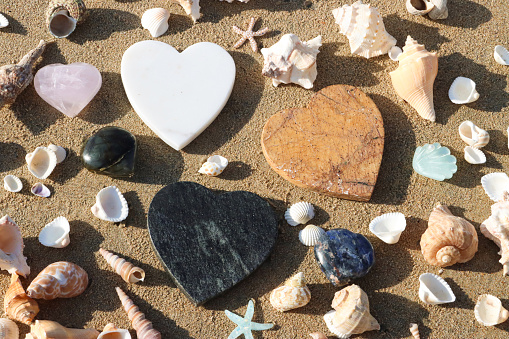 Stock photo showing close-up, elevated view of seashells with a starfish surrounding sandstone, pink quartz and granite stone hearts lying on the sand on a sunny, golden beach with sea at low tide in the background. Romantic holiday and honeymoon concept.