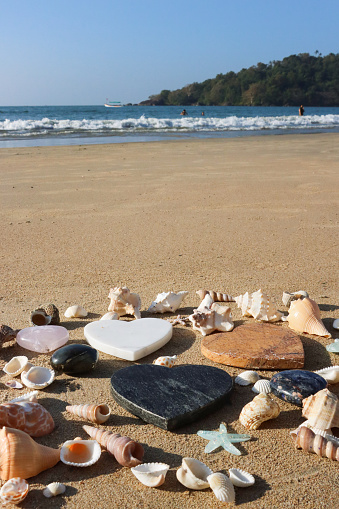 Stock photo showing close-up view of seashells with a starfish surrounding sandstone, pink quartz and granite stone hearts lying on the sand on a sunny, golden beach with sea at low tide in the background. Romantic holiday and honeymoon concept.