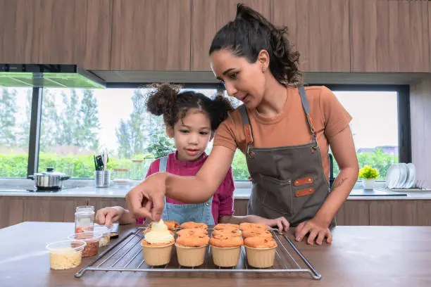 Latin American mother putting the whipped cream on the top of the bakeries while the curly-hair daughter observing and learning the bakery decoration. Happy family spending weekend time at the modern kitchen for making bakery.