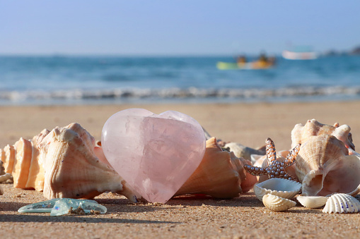 Stock photo showing close-up view of a pile of seashells with a starfish surrounding a pink quartz heart standing up in the sand on a sunny, golden beach with sea at low tide in the background. Romantic holiday and honeymoon concept.