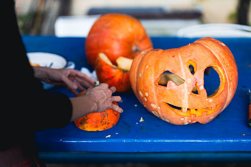 Adult Woman Hands Skillfully Carving Jack 'O' Lanterns for Upcoming Halloween Night Outdoors on a Blue Wooden Table