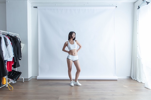 A young Asian woman in a white sports bra and shorts poses in a studio with a clothing rack in the background
