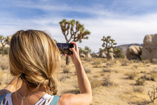 A young woman enthusiastically explores Joshua Tree National Park, capturing its beauty with her mobile phone as she hikes through the picturesque landscapes.