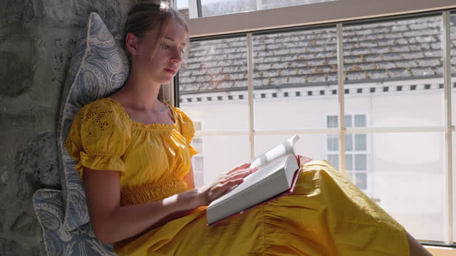 Teenage girl sitting on windowsill and reading a book