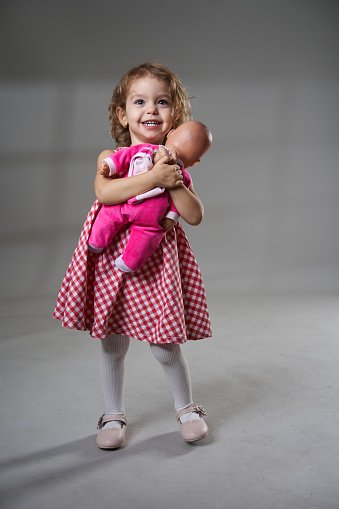 Portrait of an adorable blonde curly hair little girl holding a doll on gray background, studio shot