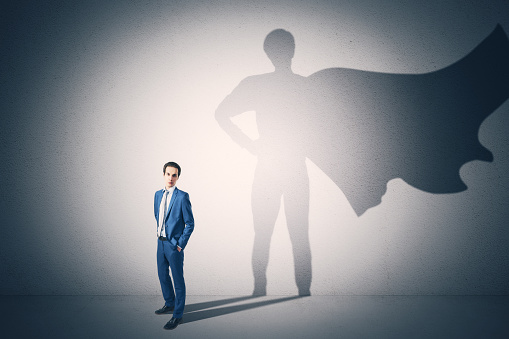 Attractive young businessman standing on concrete wall background with cape shadow. Leadership, success and power concept