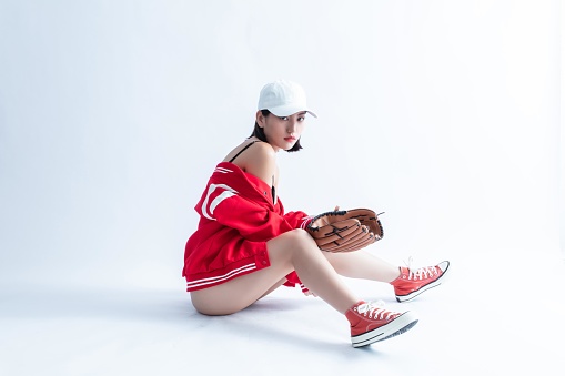 A young woman wearing a baseball cap, jacket, and sneakers is sitting on the ground and holding a baseball glove.