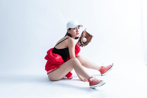 A young Asian woman wearing a baseball cap, red jacket, and sneakers is sitting on the floor and holding a baseball glove with a baseball in it.