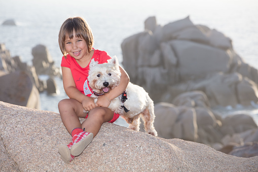 A little girl plays with her small dog among the granite boulders that form the headland opposite the Strait of Bonifacio. Both the girl and the West Highland White Terrier stick out their tongue.
