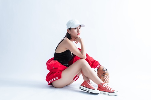 A young Asian woman wearing a baseball cap, a red jacket, and sneakers is sitting on the ground with a baseball glove and a baseball.