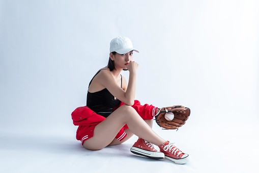 A young woman in a baseball cap and red jacket is sitting on the ground with a baseball glove and ball.