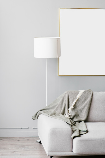 Stylish minimalistic interior of the living room in gray. Sofa with plaid, floor lamp and mockup white frame on the wall