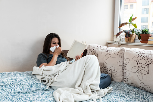 Young sick woman wrapped in a blanket wipes her nose with paper tissues, got sick because she didn't have money to pay for heating in her home or breakdown in the city heating network system.
