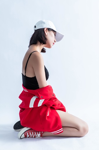 A young Asian woman wearing a baseball cap, a black sports bra, a red bomber jacket, and sneakers is sitting on the floor with her head down