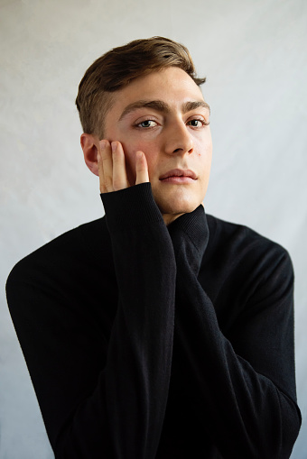 Portrait of androgynous young man on white background. He as dark blond hair and very blue eyes. He is dressed in black sweater, hands touching his face and is looking at the camera with a relaxed face. Vertical waist up studio shot in natural light. This was taken in Quebec, Canada.