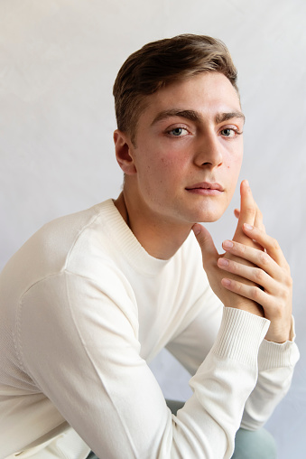 Portrait of androgynous young man on white background. He as dark blond hair and very blue eyes. He is dressed in white sweater, hands touching his face and is looking at the camera with a relaxed face. Vertical waist up studio shot in natural light. This was taken in Quebec, Canada.