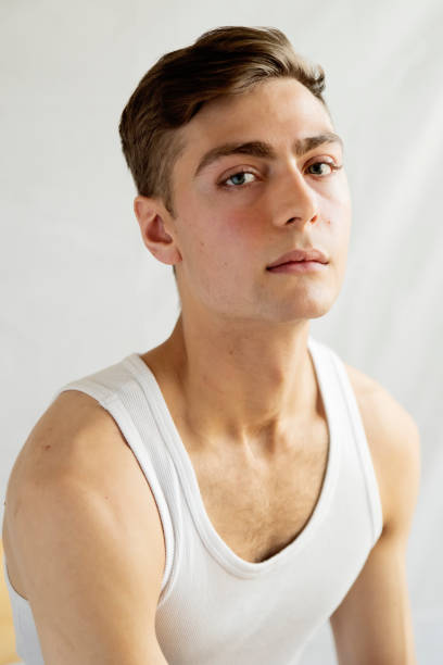 Portrait of androgynous young man on white background. He as dark blond hair and very blue eyes. He is dressed in sports clothes, white tank top and is looking at the camera with a relaxed face. Vertical waist up studio shot in natural light. This was taken in Quebec, Canada.