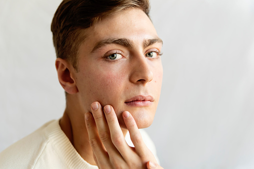Portrait of androgynous young man on white background. He as dark blond hair and very blue eyes. He is dressed in white sweater, hands touching his face and is looking at the camera with a relaxed face. Horizontal head and shoulder studio shot in natural light. This was taken in Quebec, Canada.