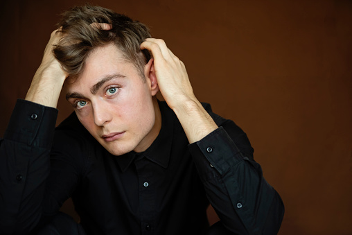 Portrait of androgynous young man on dark brown background. He as dark blond hair and very blue eyes. He is dressed in button up black shirt and is looking at the camera with a relaxed face. Horizontal waist up studio shot in natural light. This was taken in Quebec, Canada.