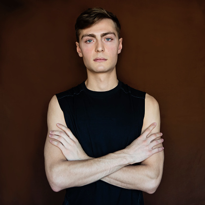 Portrait of androgynous young man on dark brown background. He as dark blond hair and very blue eyes. He is dressed in black tank top and is looking at the camera with a relaxed face. Square waist up studio shot in natural light. This was taken in Quebec, Canada.