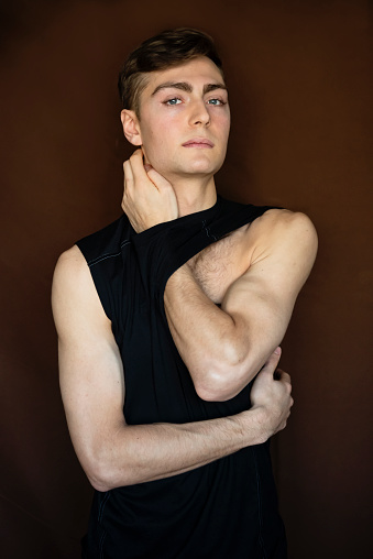 Portrait of androgynous young man on dark brown background. He as dark blond hair and very blue eyes. He is dressed in black tank top and is looking at the camera with a relaxed face. Vertical waist up studio shot in natural light. This was taken in Quebec, Canada.