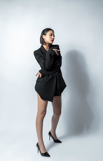 Asian woman in black suit and high heels posing in front of white background