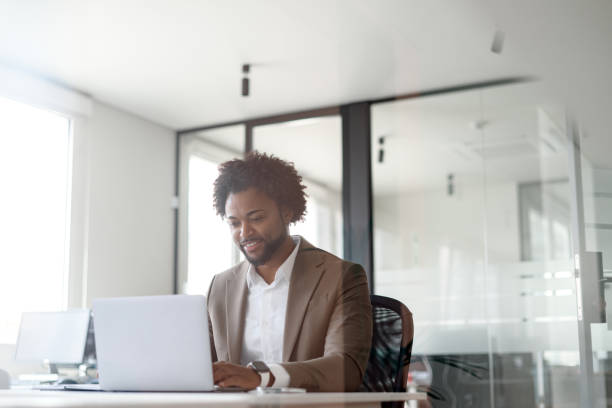 istock Smiling businessman in formal brown suit using laptop in a bright office space, conveying a concept of job satisfaction and productivity. The positive atmosphere of a well-organized work environment. 1955323347