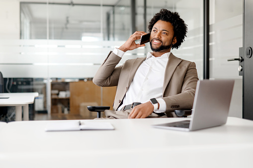 A poised African-American businessman is engaged in a phone conversation in a modern office setting, exuding confidence and professionalism. His relaxed posture and smile suggest a positive dialogue