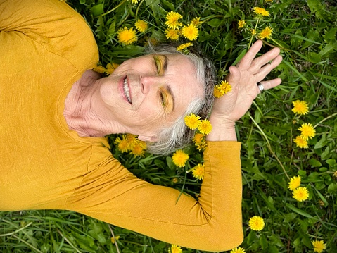 Cute and healthy senior woman, with her silver hair in a braid wrapped around her head like a crown, wearing a golden yellow shirt and yellow glittery eye make up, lying down outdoors in the long grass with lots of yellow dandelions. She is relaxing and showing vitality and aging gracefully. Her eyes are closed, enjoying the moment.