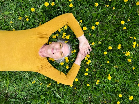 Cute and healthy senior woman, with her silver hair in a braid wrapped around her head like a crown, wearing a golden yellow shirt and yellow glittery eye make up, lying down outdoors in the long grass with lots of yellow dandelions. She is relaxing and showing vitality and aging gracefully, her arms outstretched.