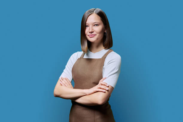 istock Young smiling woman in apron looking at camera on blue studio background 1955364514