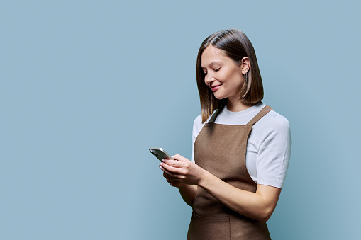 Portrait of young woman in apron holding smartphone in hands on blue background. Smiling female using mobile phone, texting receiving sending order. Technologies applications service, small business